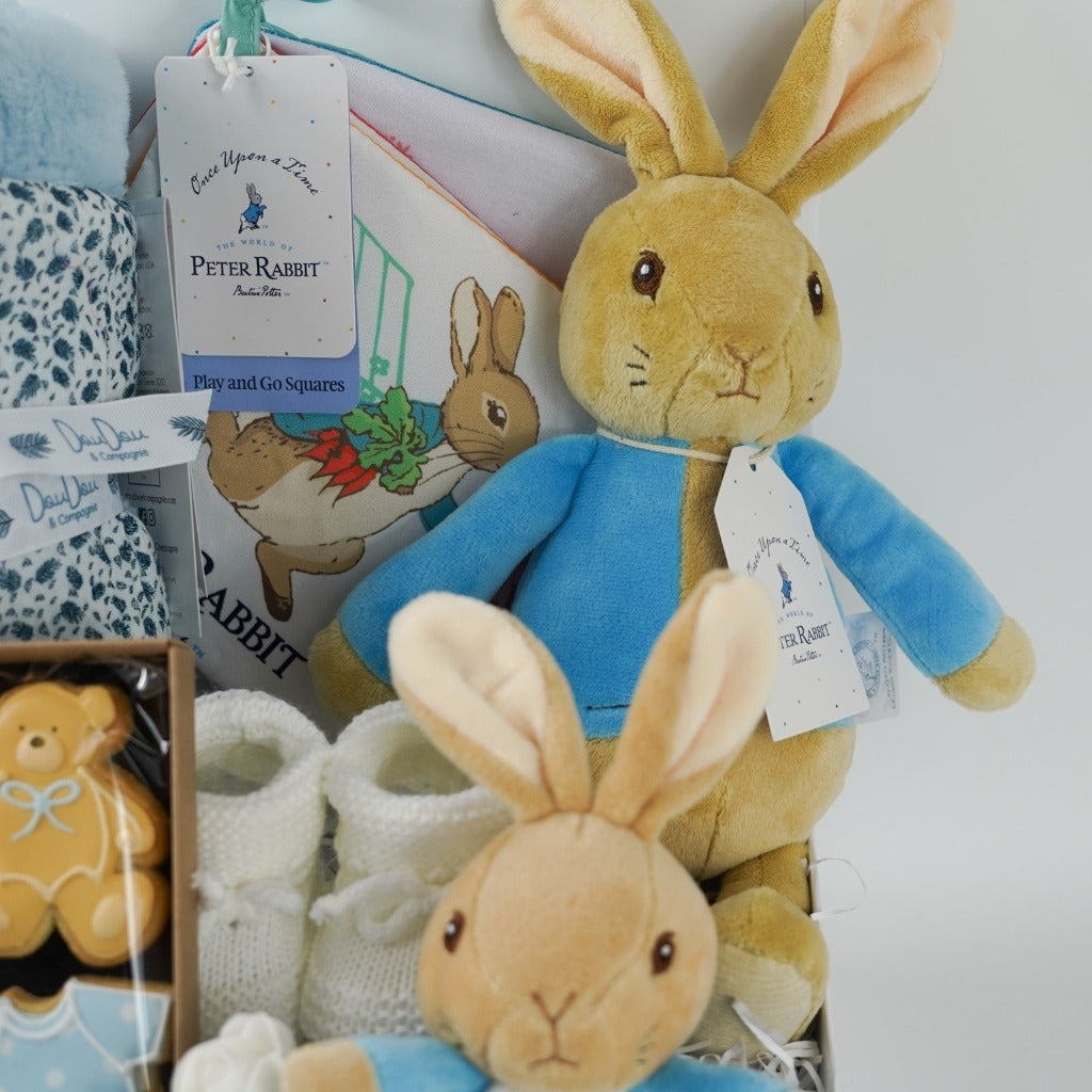 aby hamper box includes peter rabbit soft toy, Peter rabbit blue and white baby comforter, white baby knit booties, Peter rabbit cloth soft baby book, soft pale blue doudou baby blanket, baby themed celebration biscuits