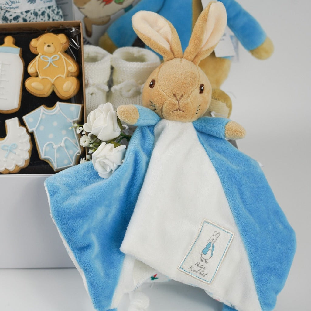 baby hamper box includes peter rabbit soft toy, Peter rabbit blue and white baby comforter, white baby knit booties, Peter rabbit cloth soft baby book, soft pale blue doudou baby blanket, baby themed celebration biscuits