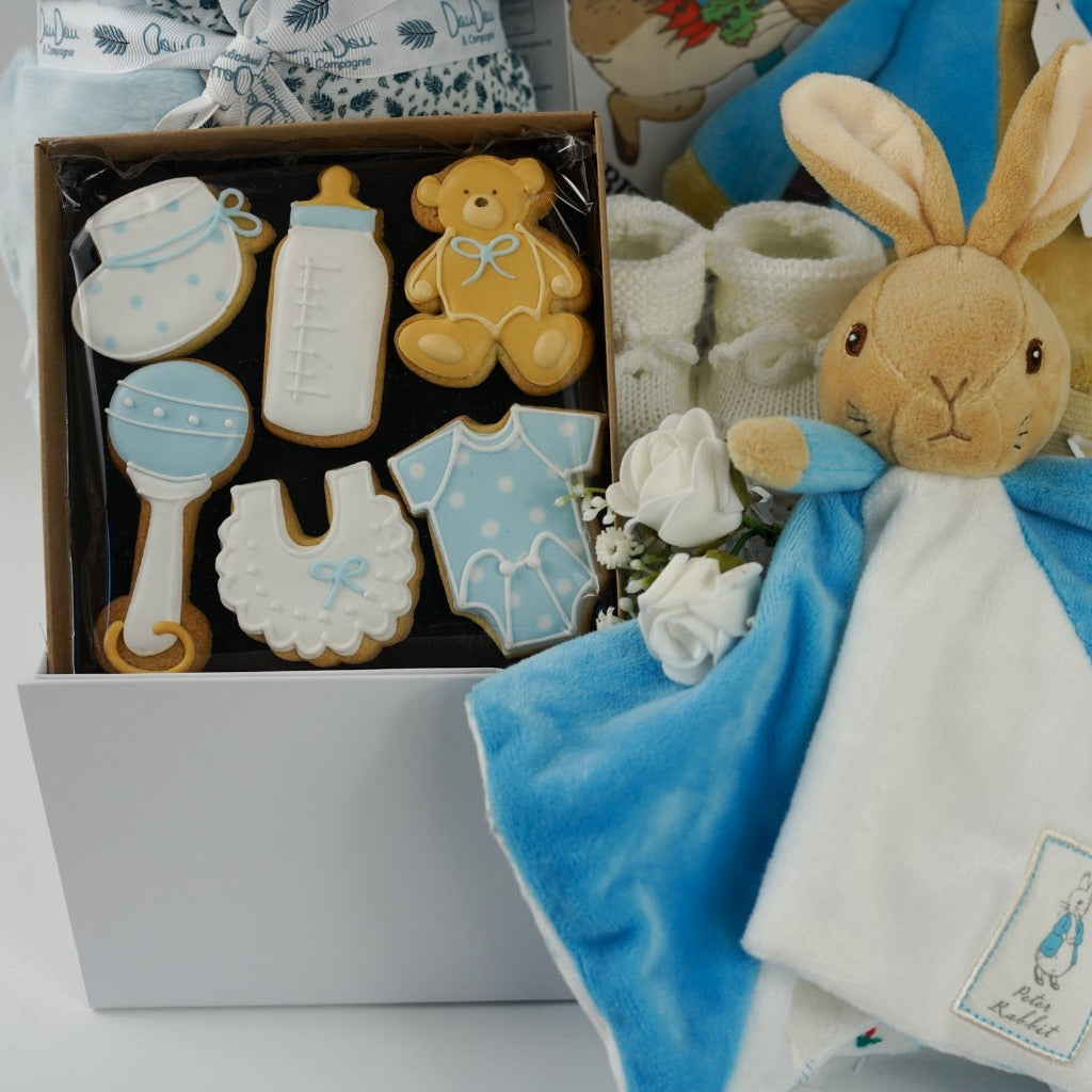 aby hamper box includes peter rabbit soft toy, Peter rabbit blue and white baby comforter, white baby knit booties, Peter rabbit cloth soft baby book, soft pale blue doudou baby blanket, baby themed celebration biscuits