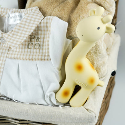 Antique washed hamper basket with Portuguese baby sleepsuit, giraffe rubber teether and bath toy in yellow, white knit bootees, white double fluffy pom pom hat, Dear mummy book, teddy comforter soft toy, butterscotch coloured dressing gown with ears