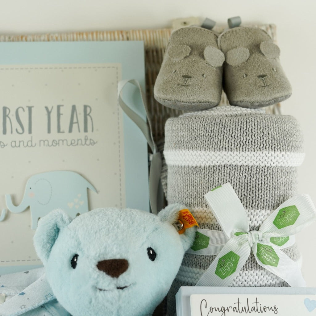 Luxury baby hamper in a wicker basket, Blue steiff my first teddy, Star and Crown baby sleepsuit in blue, My First year blue baby book, grey baby suede look slippers with cute face, grey and white heave baby blanket, forever baby sentiment card in blue with elephant design