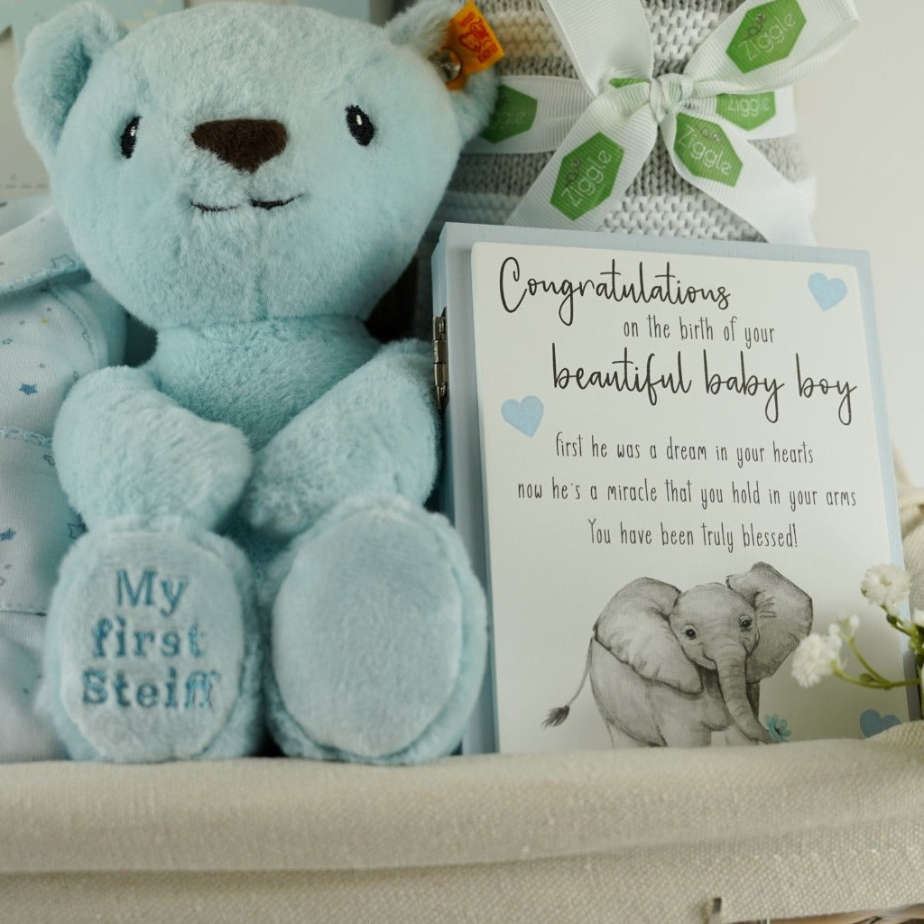 Luxury baby hamper in a wicker basket, Blue steiff my first teddy, Star and Crown baby sleepsuit in blue, My First year blue baby book, grey baby suede look slippers with cute face, grey and white heave baby blanket, forever baby sentiment card in blue with elephant design