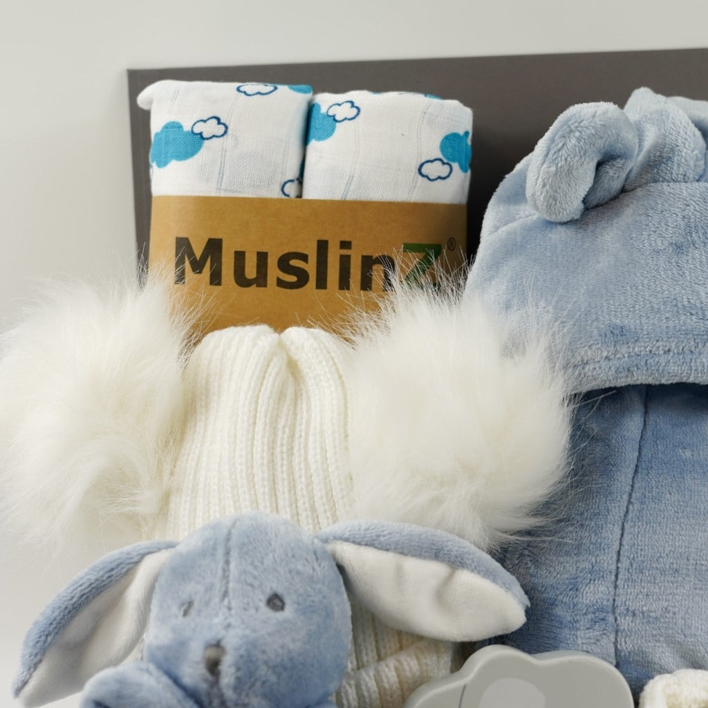 Grey magnetic box with dusty blue baby dressing gown with cute ears, dusty blue baby rabbit comforter, white baby bobble hat with 2 fluffy pom poms, grey wooden push along elephant toy, two white organic muslins with blue cloud pattern, white knit baby bootees