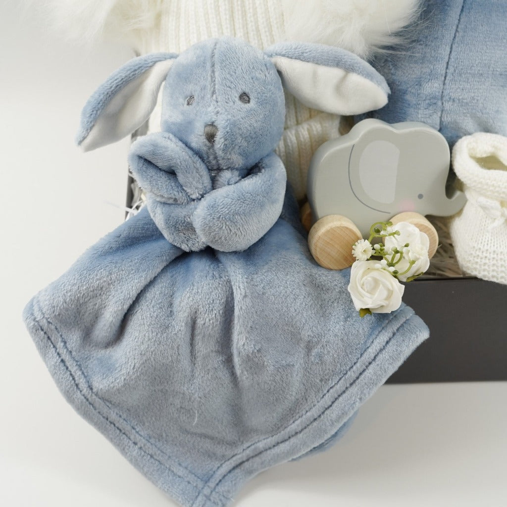 Grey magnetic box with dusty blue baby dressing gown with cute ears, dusty blue baby rabbit comforter, white baby bobble hat with 2 fluffy pom poms, grey wooden push along elephant toy, two white organic muslins with blue cloud pattern, white knit baby bootees