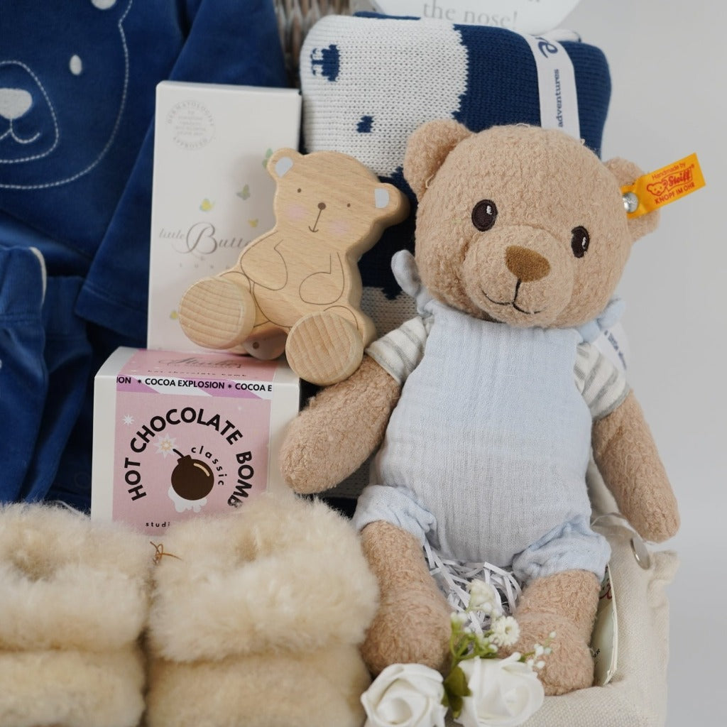 baby boy hamper with blue velour organic top and leggings, alpaca baby slippers, steiff organic bear, organic blue and white teddy bear blanket , wooden bear poush along toy , natural baby toiletries in a white wicker hamper basket