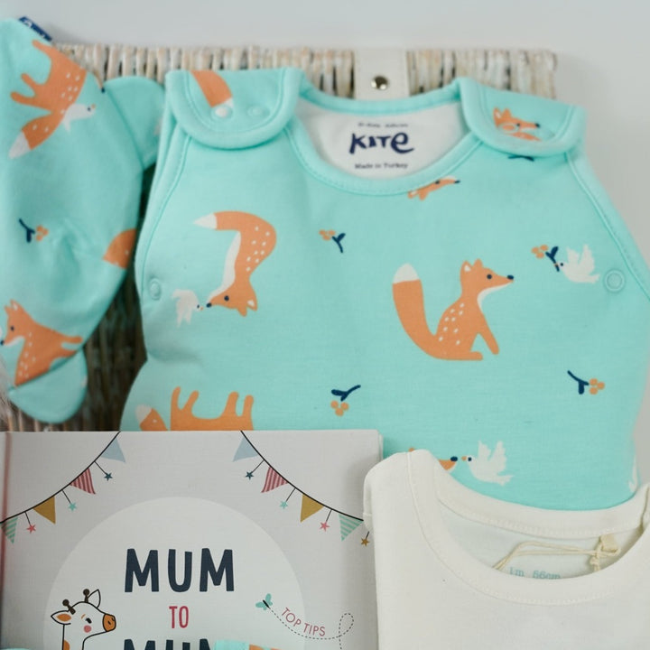 mint green baby sleepsuit with foxes and doves, baby sleeping bag with foxes and doves, baby organic cotton hat with foxes and doves, baby vest in white with foxes and doves, mum to mum advice book, white knit booties, coral coloured soft fox toy in a white hamper basket