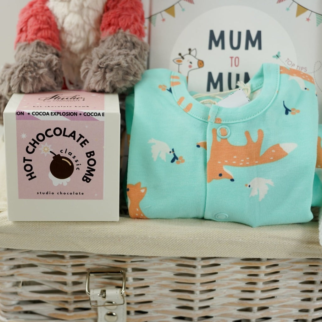 mint green baby sleepsuit with foxes and doves, baby sleeping bag with foxes and doves, baby organic cotton hat with foxes and doves, baby vest in white with foxes and doves, mum to mum advice book, white knit booties, coral coloured soft fox toy in a white hamper basket