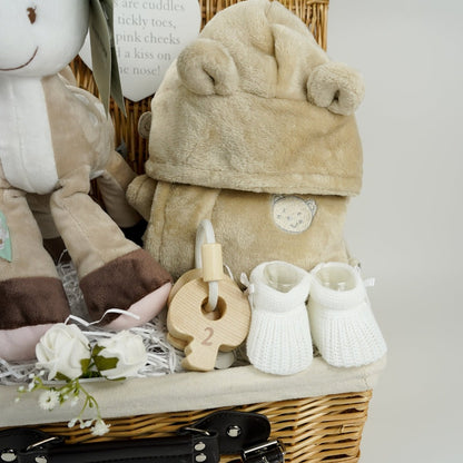 baby hamper in wicker basket with a fawn soft toy, white cotton cellular blanket, while soft large muslin with grey elephants and stars, soft fawn coloured dressing gown with cute teddy ears, white knit booties, set of wooden teething keys on a rope, hot chocolate bomb