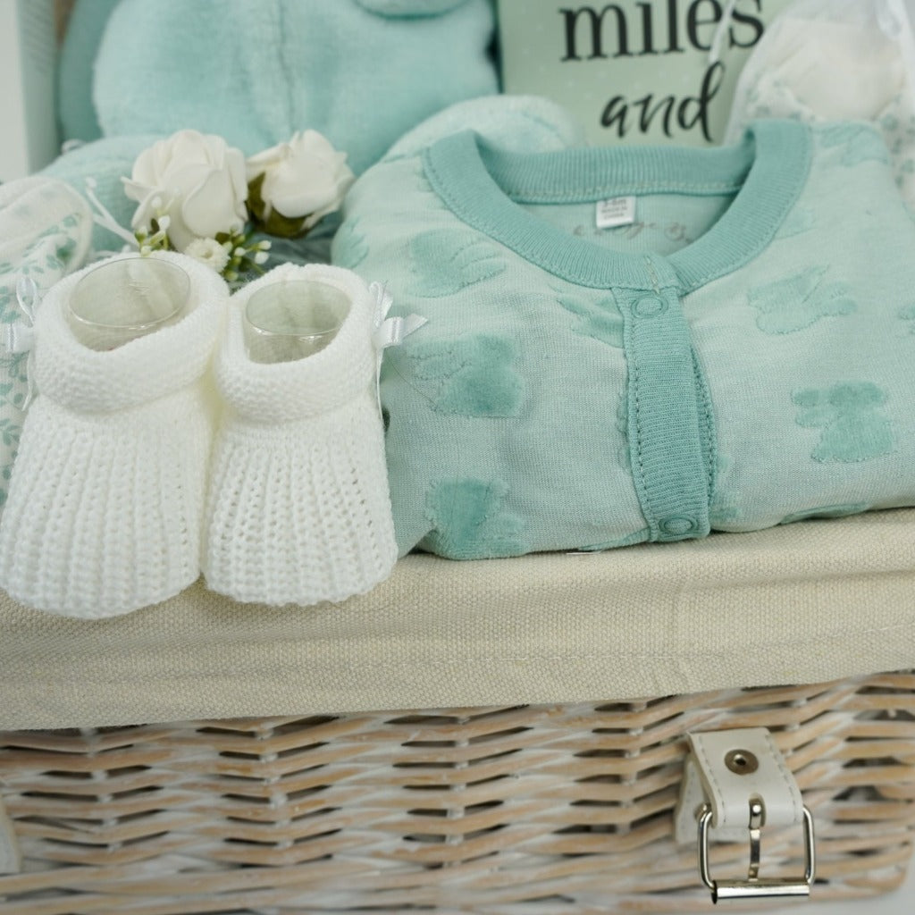 white hamper basket with mint green baby bunny soft toy by nattou, baby toiletries including bubble bath and nappy cream, green baby bunny layetter, bunny swaddle , boxed miles and moments baby milestone cards, white knit booties