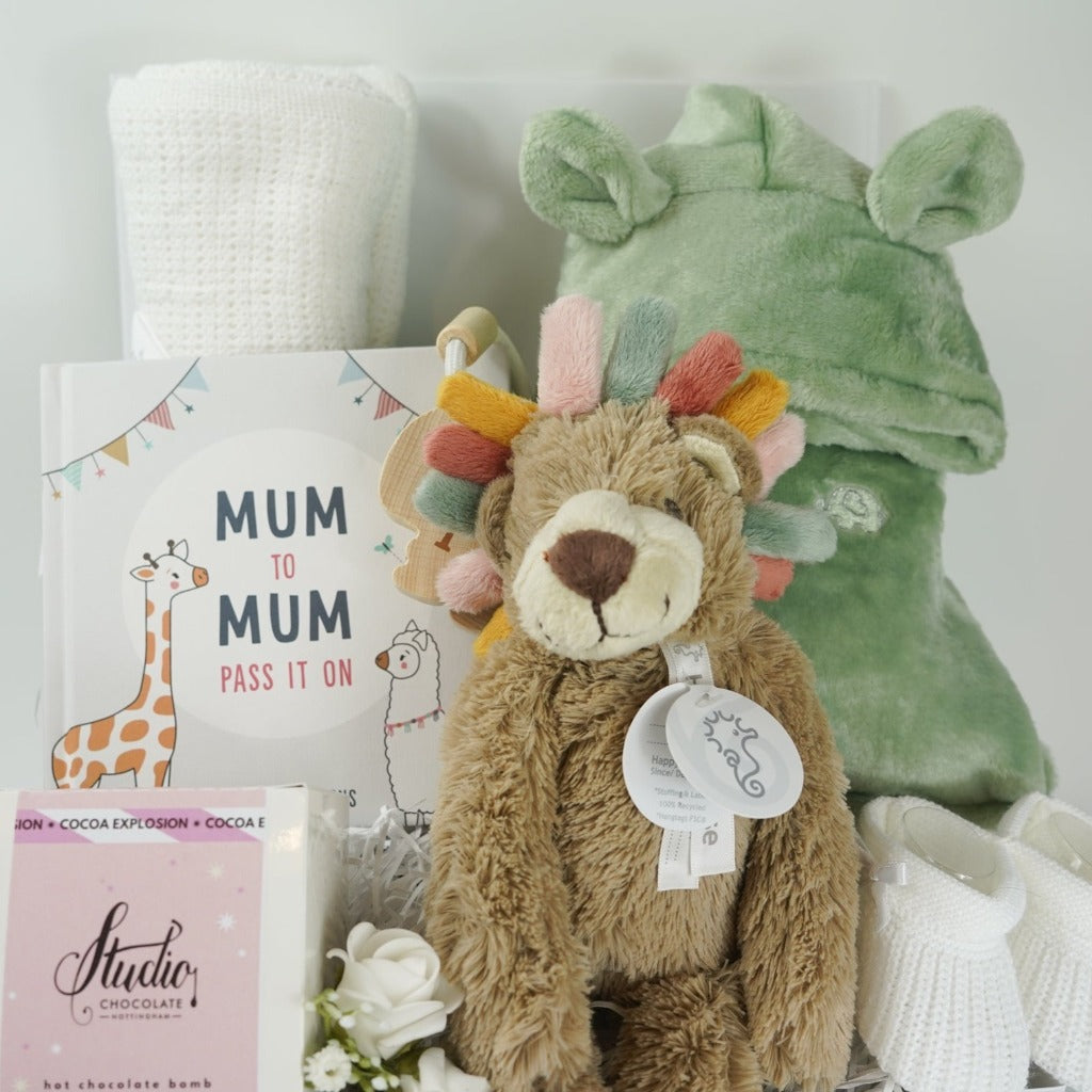 White hamper box with white cellular baby blanket, sage green fleece baby dressing gown with cute ears , Lion teddy with colourful taggie mane, hot chocolate bomb, white knit baby booties, Sunshine wooden nursery plaque
