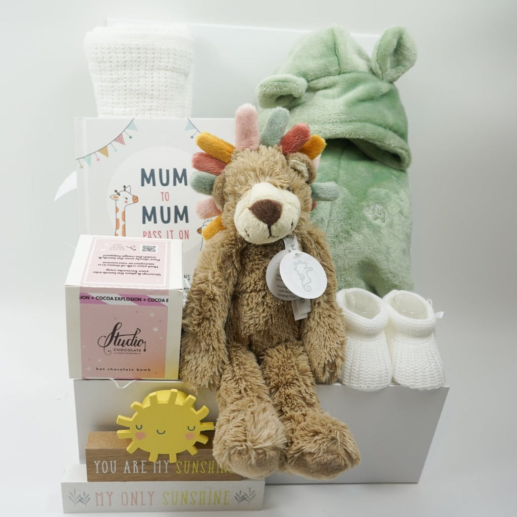 White hamper box with white cellular baby blanket, sage green fleece baby dressing gown with cute ears , Lion teddy with colourful taggie mane,  hot chocolate bomb, white knit baby booties, Sunshine wooden nursery plaque