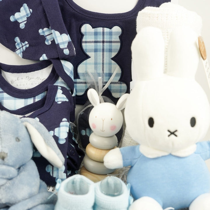 baby boy hamper includes navy baby layette with pale blue tartan teddy design includes a sleepsuit, bodysuit, hat, mittens and bib, white cotton cellular blanket, wooden rabbit stacking toy, dusky blue rabbit comforter, blue knit booties, My First Miffy white toy with a blue outfit