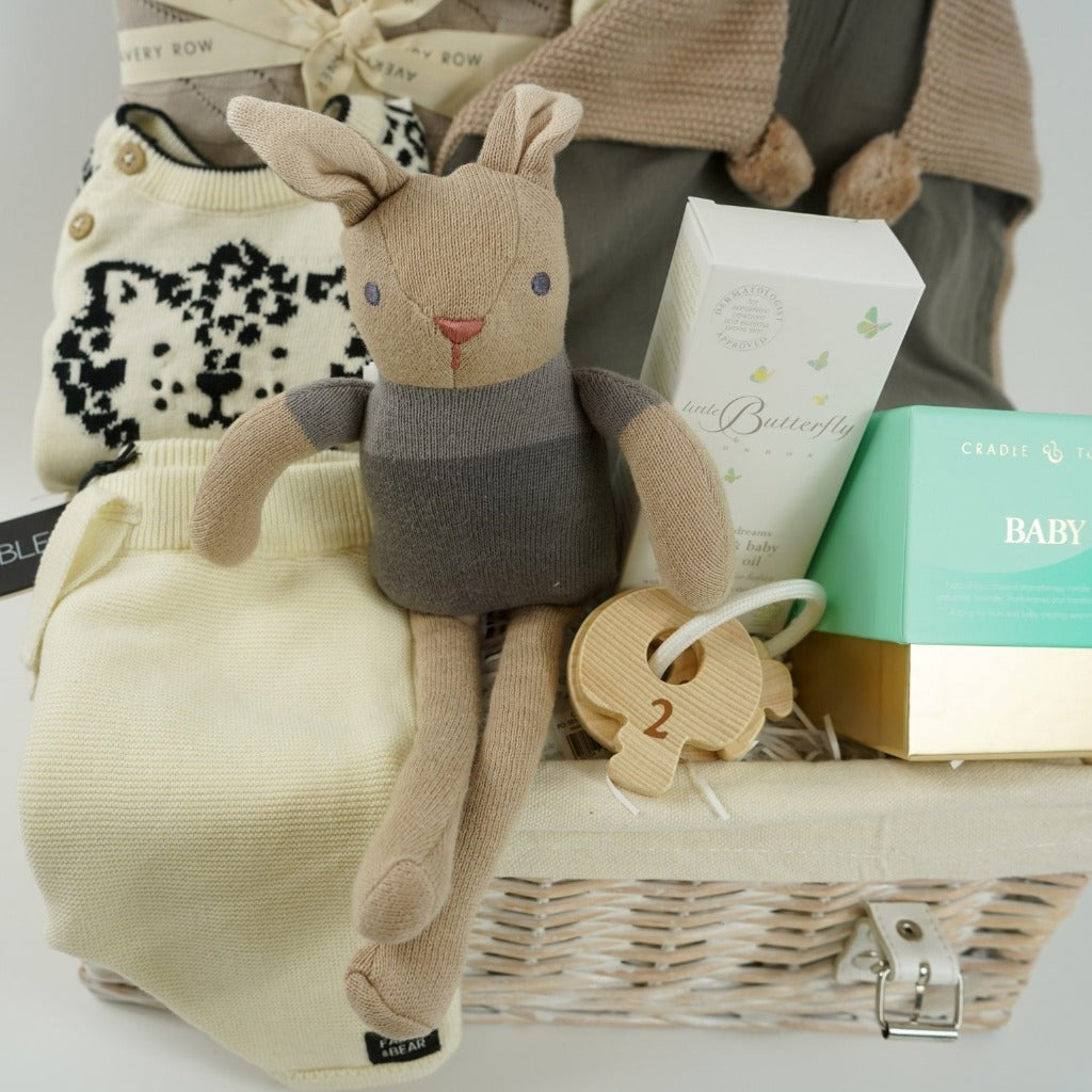 White wicker hamper basket, organic baby rabbit toy and matching comforter, Knitted organic baby clothing set including a jumper with leopard design, cradle and tonic baby candle, organic baby massage oil, wooden teething keys