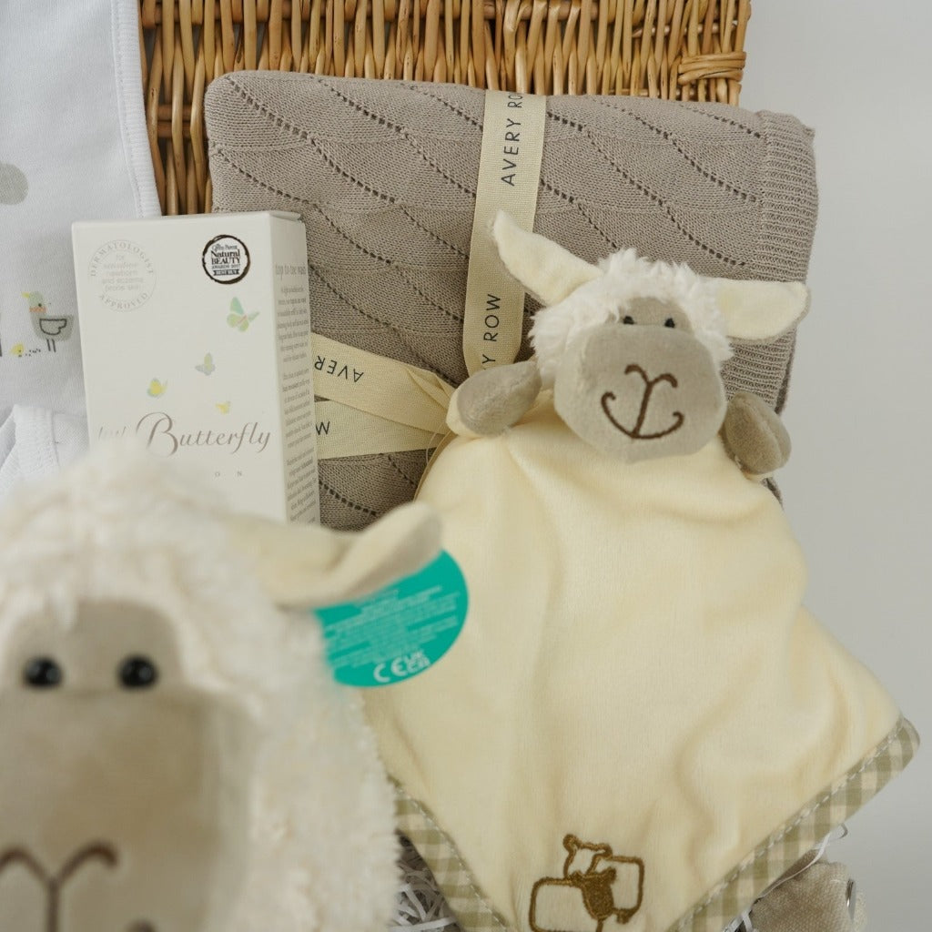 wicker hamper basket with white cotton baby clothing set with sheep design, organic baby blanket in buscuit colour, sheep soft toy, sheep rattle and sheep baby comforter finger puppet, wooden baby teether, organic baby wash