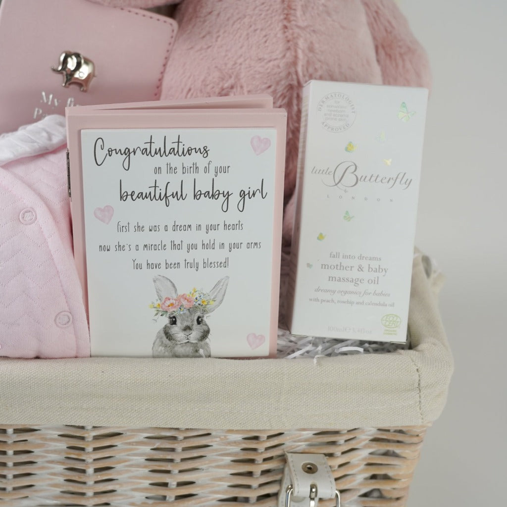 White hamper basket with baby girl gifts including pink baby quilted outfit with white frilled collar, baby pink passport holder and luggage label with silver elephant, pink and white heavy knit baby blanket, pink soft cuddly eco friendly bunny, little butterfly london organic baby and mum massage oil in a box, wooden forever congratulation card
