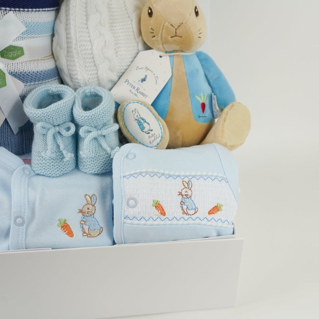 baby hamper with peter rabbit blue outfit including a pale blue sleepsuit and matching jacket, pale blue booties, blue and white and beige baby blanket, white pom pom hat and peter rabbit soft toy