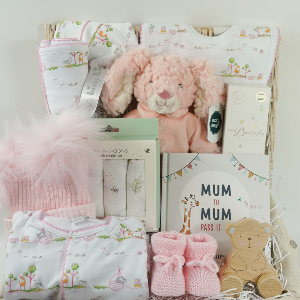 white hamper basket with Easter gifts for a baby girl, pink bunny in pink pjamas, Kissy Kissy Noahs ark baby clothing set in prima cotton includes white with pink trink sleepsuit, bib and hat, Noah's Ark pattern pink and white baby blanket, baby wash cloths with bunnies, Organic baby wash, pink knit booties, wooden teddy, pink fluffy double pom pom baby hat, mum to mum book full of tips