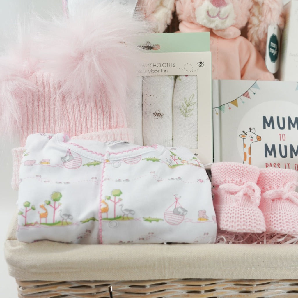 white hamper basket with Easter gifts for a baby girl, pink bunny in pink pjamas, Kissy Kissy Noahs ark baby clothing set in prima cotton includes white with pink trink sleepsuit, bib and hat, Noah's Ark pattern pink and white baby blanket, baby wash cloths with bunnies, Organic baby wash, pink knit booties, wooden teddy, pink fluffy double pom pom baby hat, mum to mum book full of tips