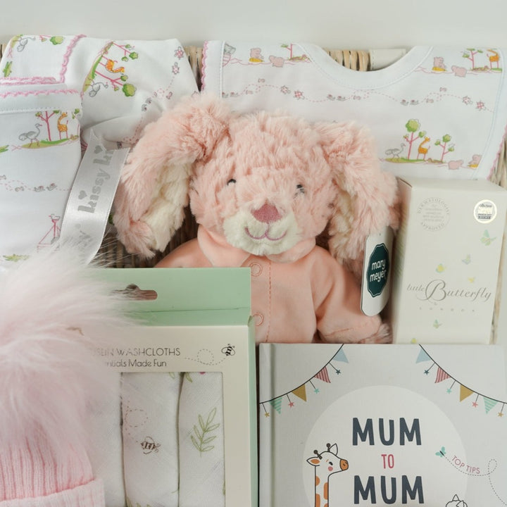 white hamper basket with Easter gifts for a baby girl, pink bunny in pink pjamas, Kissy Kissy Noahs ark baby clothing set in prima cotton includes white with pink trink sleepsuit, bib and hat, Noah's Ark pattern pink and white baby blanket, baby wash cloths with bunnies, Organic baby wash, pink knit booties, wooden teddy, pink fluffy double pom pom baby hat, mum to mum book full of tips