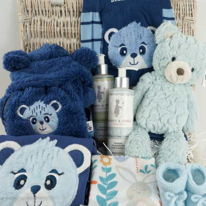 white wicker hamper basket, blue with teddy design baby clothing set includes baby leggings with teddy on the bum, feece jacket with hood in navy with teddy and blue long sleeved top with teddy face, organic toiletries, pale blue teddy, swaddle with teddy and owl design, pale blue knit booties