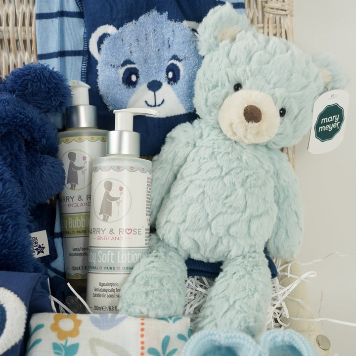 white wicker hamper basket, blue with teddy design baby clothing set includes baby leggings with teddy on the bum, feece jacket with hood in navy with teddy and blue long sleeved top with teddy face, organic toiletries, pale blue teddy, swaddle with teddy and owl design, pale blue knit booties