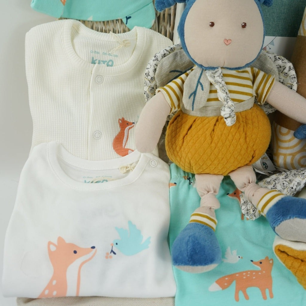 Baby hamper with organic clothing set includes baby sleepsuit in white with fox and dove design, baby bodysuit with long sleeves and fox and dove design, baby leggings with fox and matching hat, Organic cotton bee soft toy and matching comforter, organic cotton white swaddles with yellow design, green baby journal, organic baby toiletries