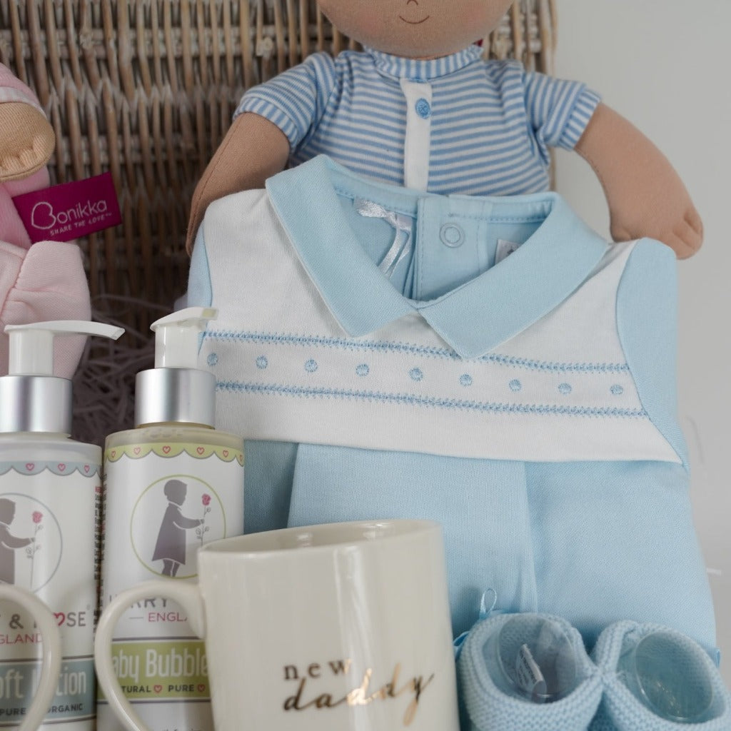 Twin baby hamper includes pink luxury baby girl outfit with a white collar and bows, blue baby boy outfit with a blue collar and white yolk, pink soft baby ragdool with a pink hat, blue soft baby ragdoll with sriped blue and white outfit and blue hat, pink booties and blue booties, organic baby lotion and organic bubble bath , new mummy and daddy cream bone china mug