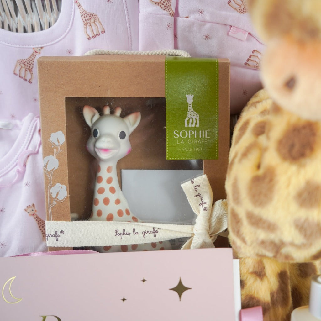 White hamper basket with baby girl gifts, luxury pink sophie la girafe baby outfit with frill at the front, includes sleepsuit, hat and bib, sophie la girafe teething toy, big soft floppy girafe eco friendly toy, pink baby shoes in suede effect with cute ears and face, pink baby photo album , baby toiletries