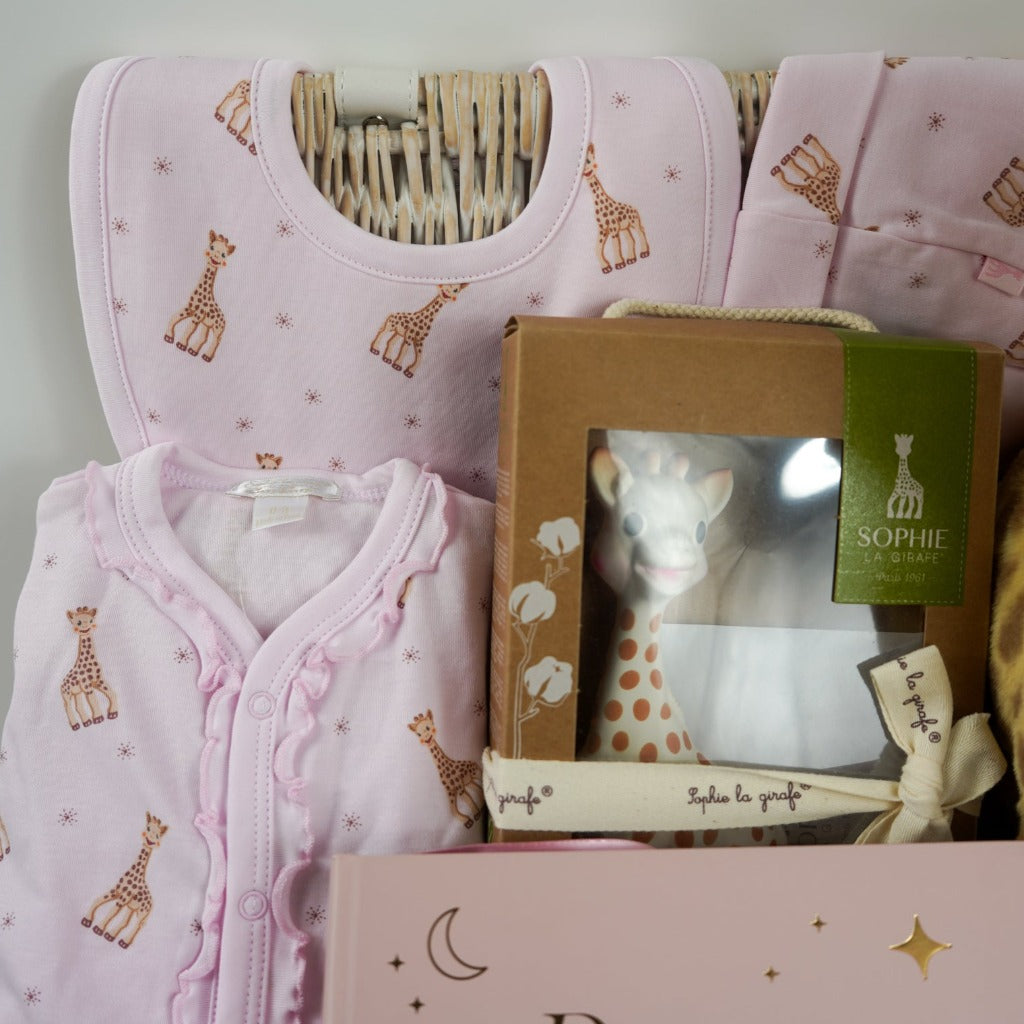 White hamper basket with baby girl gifts, luxury pink sophie la girafe baby outfit with frill at the front, includes sleepsuit, hat and bib, sophie la girafe teething toy, big soft floppy girafe eco friendly toy, pink baby shoes in suede effect with cute ears and face, pink baby photo album , baby toiletries