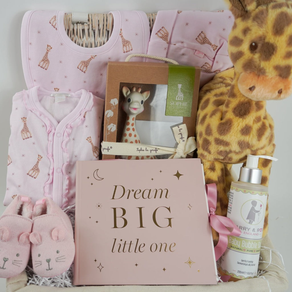 White hamper basket with baby girl gifts, luxury pink sophie la girafe baby outfit with frill at the front, includes sleepsuit, hat and bib, sophie la girafe teething toy, big soft floppy girafe eco friendly toy, pink baby shoes in suede effect with cute ears and face, pink baby photo album , baby toiletries