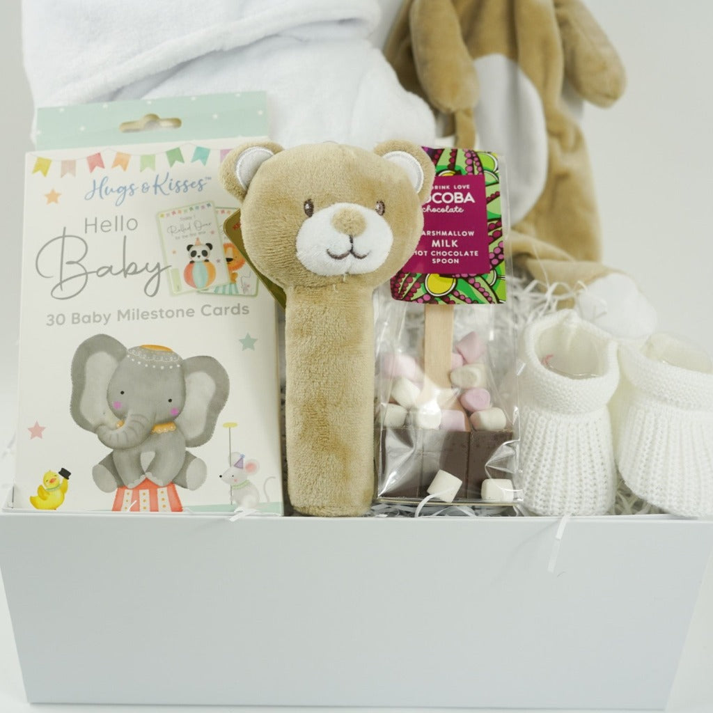 white hamper box with baby items including baby dressing gown in white with cute ears, soft brown baby comforter and matching squeaky rattle, baby milestone cards with cute animals, white knit baby booties , hot chocolate spoon