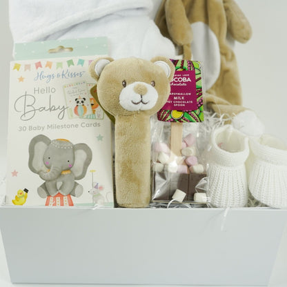 white hamper box with baby items including baby dressing gown in white with cute ears, soft brown baby comforter and matching squeaky rattle, baby milestone cards with cute animals, white knit baby booties , hot chocolate spoon
