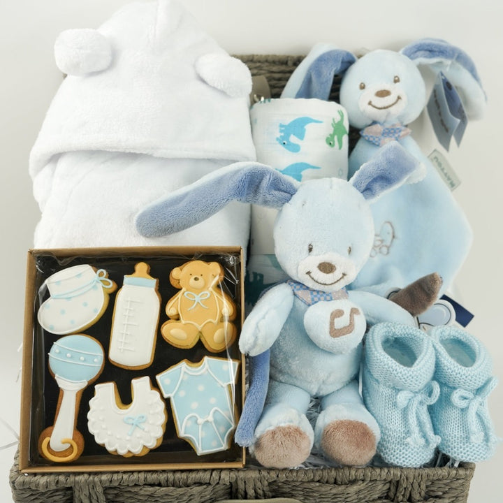 Baby boy hamper basket, white baby dressing gown with ears, muslin with dinosaurs in green and blue, blue Nattou rabbit musicaql and matching comforter, baby booties in blue, box of baby themed biscuits in blue and white