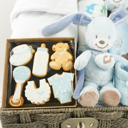 Baby boy hamper basket, white baby dressing gown with ears, muslin with dinosaurs in green and blue, blue Nattou rabbit musicaql and matching comforter, baby booties in blue, box of baby themed biscuits in blue and white