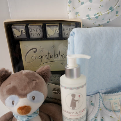 baby boy hamper box, newborn baby bodysuit with long sleeves and matching baby hat, blue baby wrap with satin ribbon edging, musical racoon soft toy, congratulations chocolates, white knit booties and baby body lotion