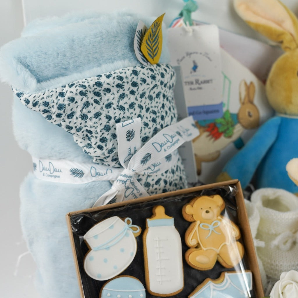 aby hamper box includes peter rabbit soft toy, Peter rabbit blue and white baby comforter, white baby knit booties, Peter rabbit cloth soft baby book, soft pale blue doudou baby blanket, baby themed celebration biscuits