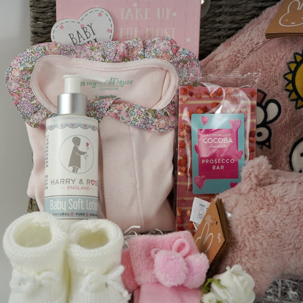 Grey baby hamper box with pink magnetic fastening baby sleepsuit with frilled liberty collar, baby booties, pink snuffie miffy and friends soft dog, soft pink fluffy book, prosecco chocolate bar, organic baby lotion, baby socks in pink with a slogan on the foot and pom poms , white knit booties