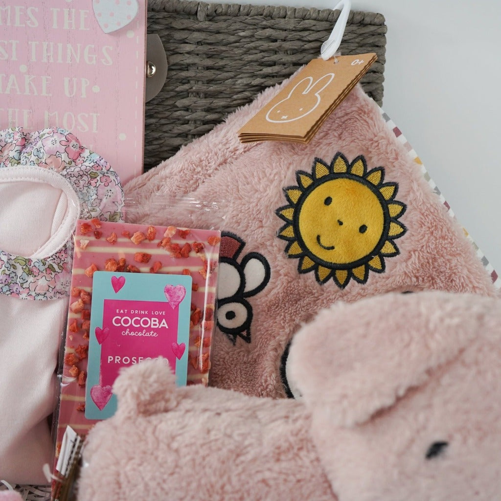 Grey baby hamper box with pink magnetic fastening baby sleepsuit with frilled liberty collar, baby booties, pink snuffie miffy and friends soft dog, soft pink fluffy book, prosecco chocolate bar, organic baby lotion, baby socks in pink with a slogan on the foot and pom poms , white knit booties