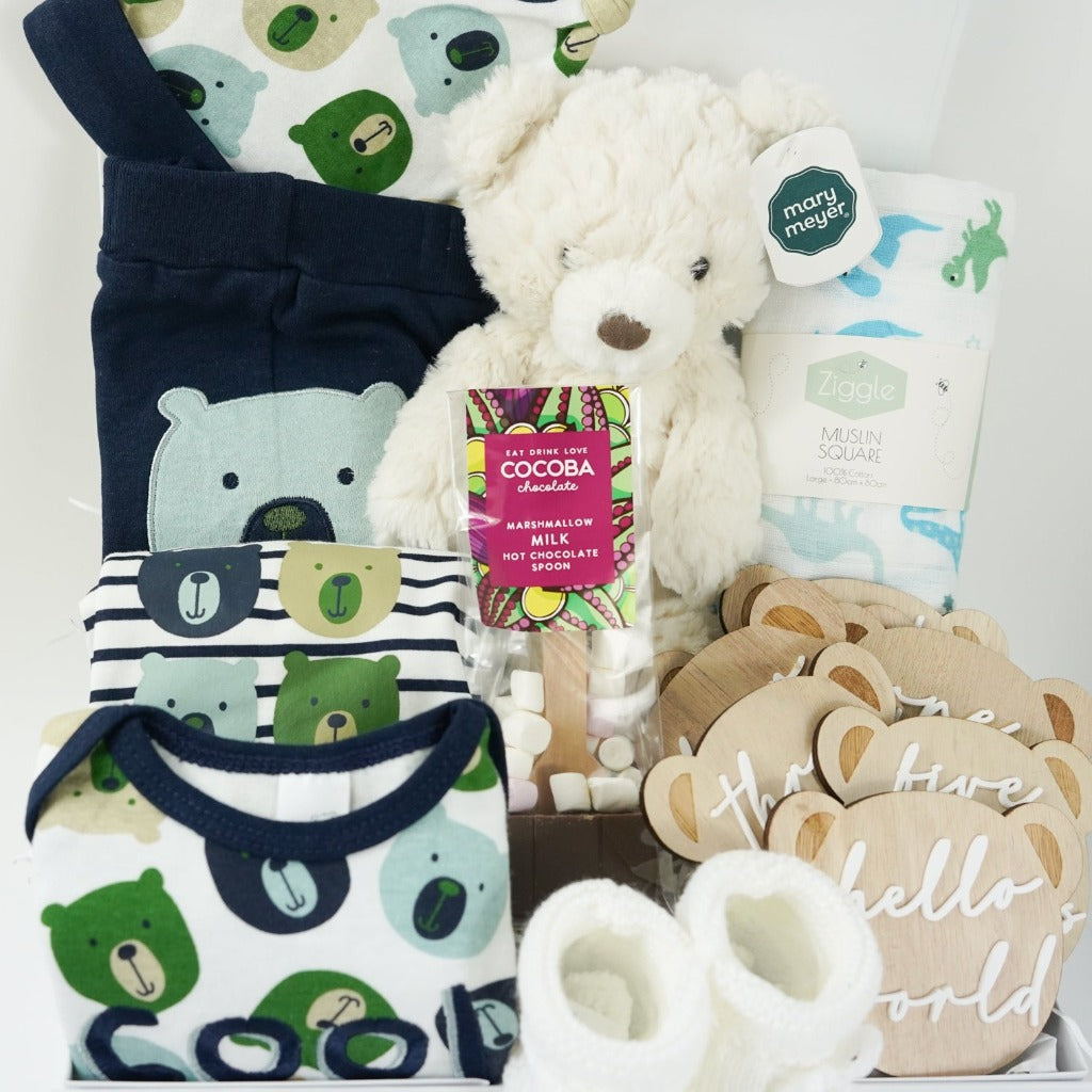 baby hamper with navy blue baby set including baby joggers and 2 baby vests and match hat, milestone wooden teddy discs , cocoba chocolate spoon with marshmallows, baby white knit booties, cream teddy, white muslin weith blue and green dinosaurs