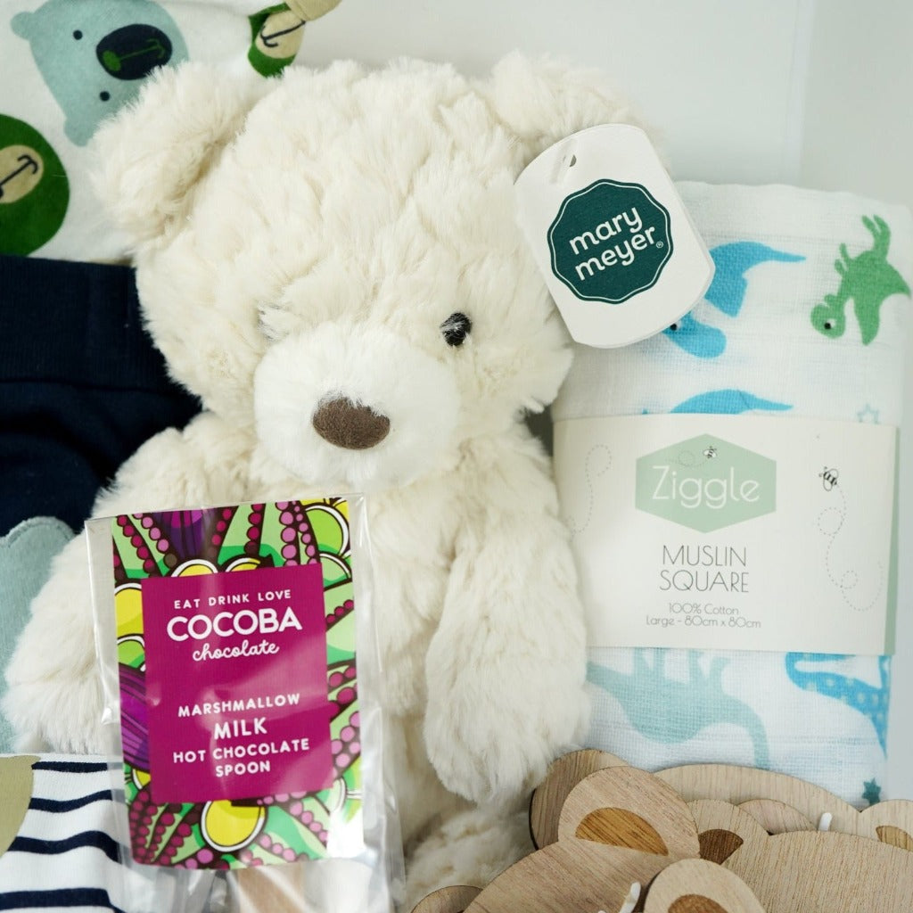 baby hamper with navy blue baby set including baby joggers and 2 baby vests and match hat, milestone wooden teddy discs , cocoba chocolate spoon with marshmallows, baby white knit booties, cream teddy, white muslin with blue and green dinosaurs