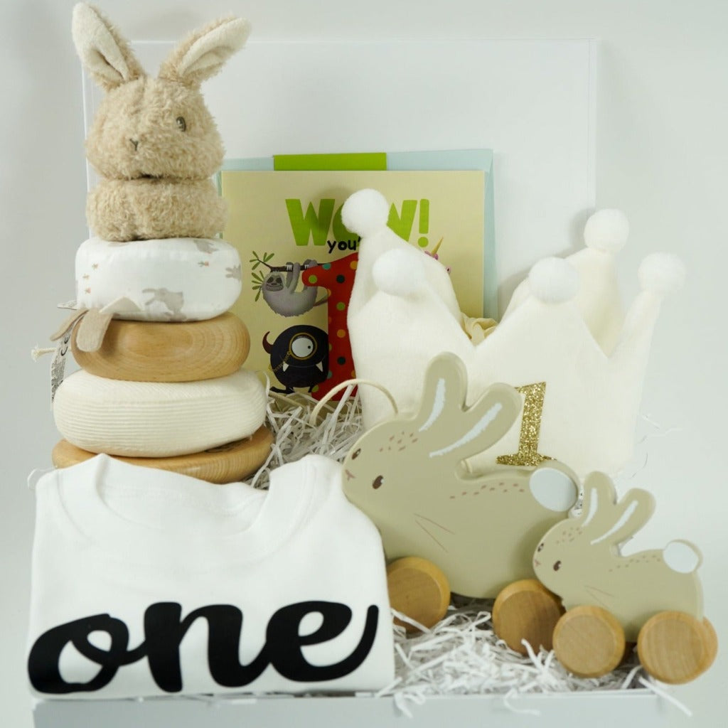 First birthday hamper contains wooden bunny stacker, 2 wooden pull along bunnies, white t shirt with one on it, white crown with pom poms and number 1 