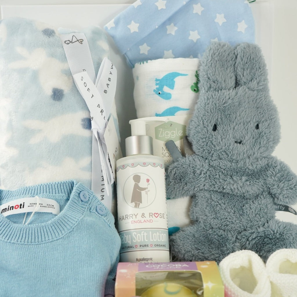 baby boy hamper box with blue and white striped fine knit baby romper, blue baby blanket with white bunnies, white muslin with blue and green dinosaurs, fluffy blue miffy comforter with dummy tag, white knit booties, organic baby soft lotion in a pump dispenser, white hot chocolate bombe