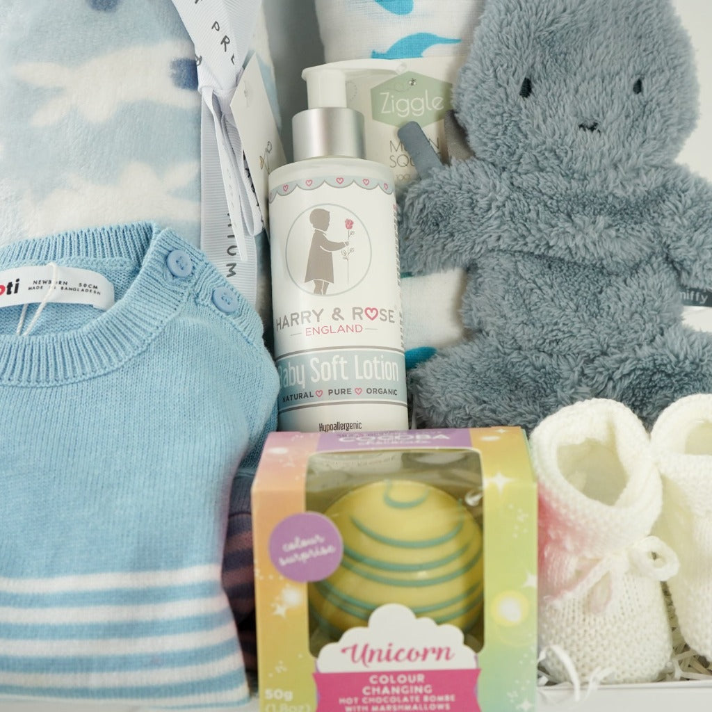 baby boy hamper box with blue and white striped fine knit baby romper, blue baby blanket with white bunnies, white muslin with blue and green dinosaurs, fluffy blue miffy comforter with dummy tag, white knit booties, organic baby soft lotion in a pump dispenser, white hot chocolate bombe