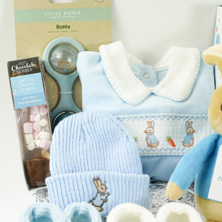 white baby hamper box with Beatrix potter Peter rabbit soft toy, blue velour baby sleepsuit with smocked yolk with bunny's , blue beanie hat with bunny, Little Dutch baby rattle, white muslin swaddle with blue elephants, salted caramel chocolate spoon with marshmallow