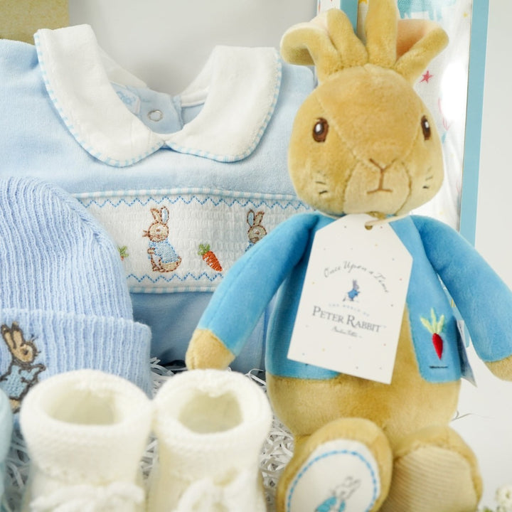 white baby hamper box with Beatrix potter Peter rabbit soft toy, blue velour baby sleepsuit with smocked yolk with bunny's , blue beanie hat with bunny, Little Dutch baby rattle, white muslin swaddle with blue elephants, salted caramel chocolate spoon with marshmallow