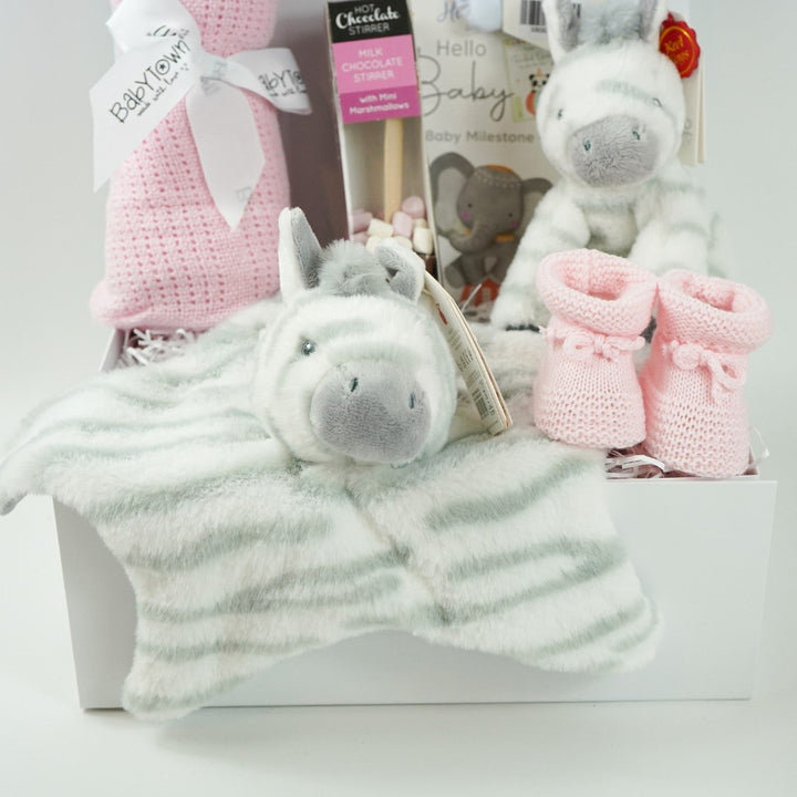 White hamper box with pink cellular baby blanket, white knot hat with silver stars, baby milestone cards, chocolate stirrer spoon with marshmallows, pink knit baby booties , grey and white zebra soft toy and matching comforter