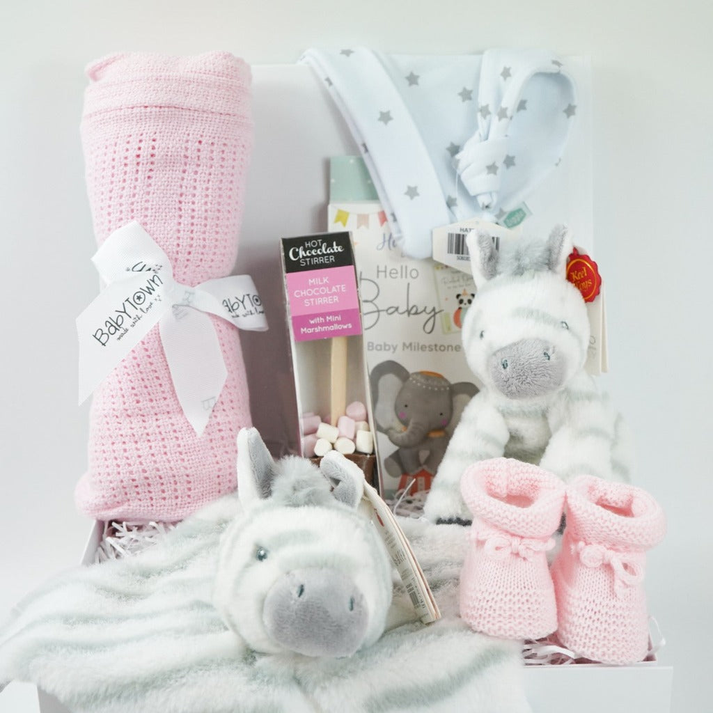 White hamper box with pink cellular baby blanket, white knot hat with silver stars, baby milestone cards, chocolate stirrer spoon with marshmallows, pink knit baby booties , grey and white zebra soft toy and matching comforter