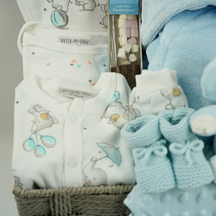 baby boy hamper gift includes white baby layette with grey elephants with blue accents, blue baby dressing gown, blue elephant soft, blue knit booties, chocolate stirring spoon, baby taggie comfort blanket