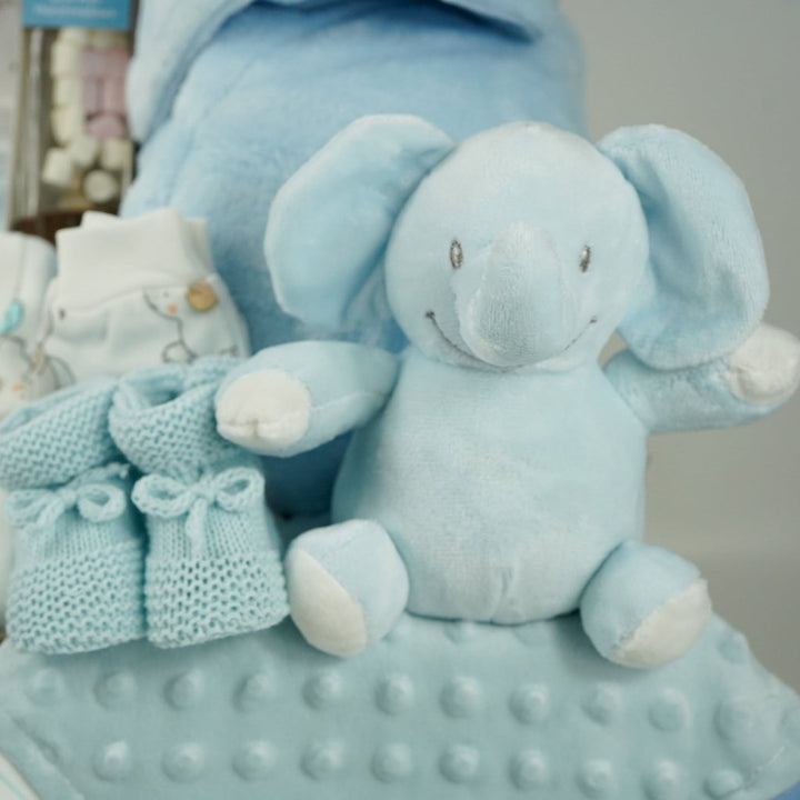 baby boy hamper gift includes white baby layette with grey elephants with blue accents, blue baby dressing gown, blue elephant soft, blue knit booties, chocolate stirring spoon, baby taggie comfort blanket