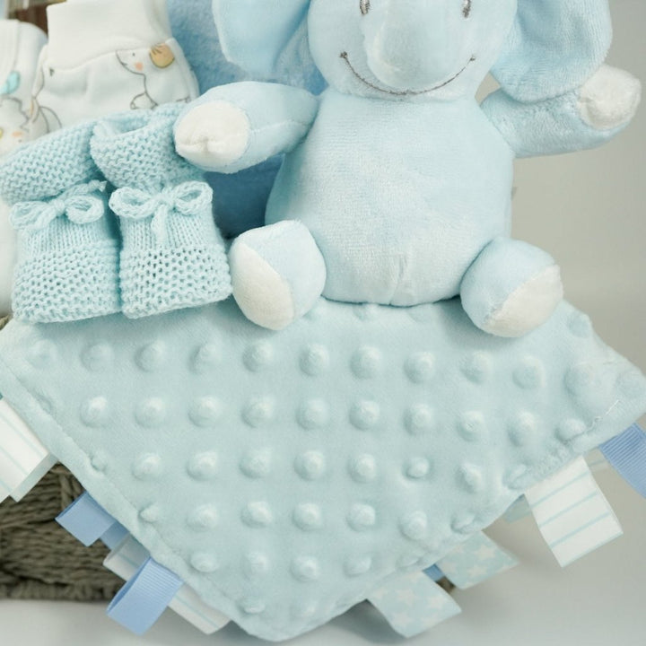 baby boy hamper gift includes white baby layette with grey elephants with blue accents, blue baby dressing gown, blue elephant soft, blue knit booties, chocolate stirring spoon, baby taggie comfort blanket